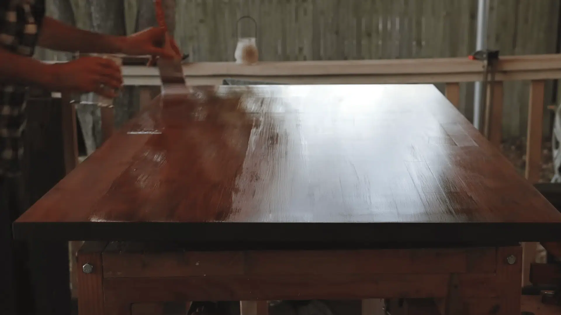 a person brushes finish onto a large dark wooden tabletop resting on a workbench on an outdoor porch