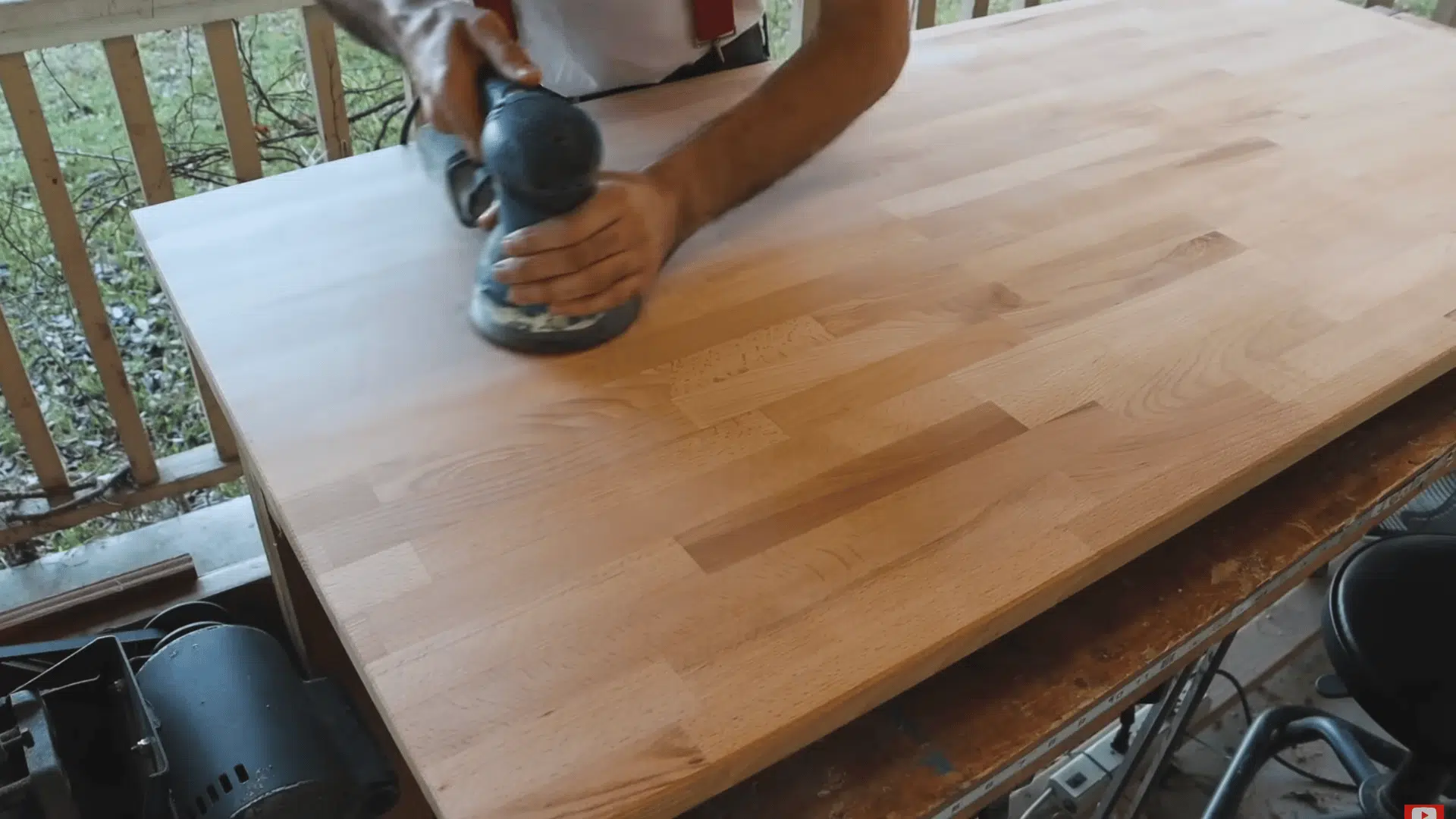 a person sands a large wooden tabletop with an orbital sander on an outdoor workbench near a porch railing