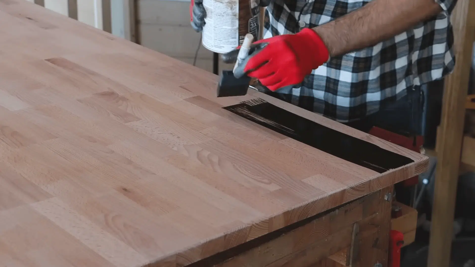 a person wearing red gloves applies dark wood stain to the edge of a tabletop with a foam brush