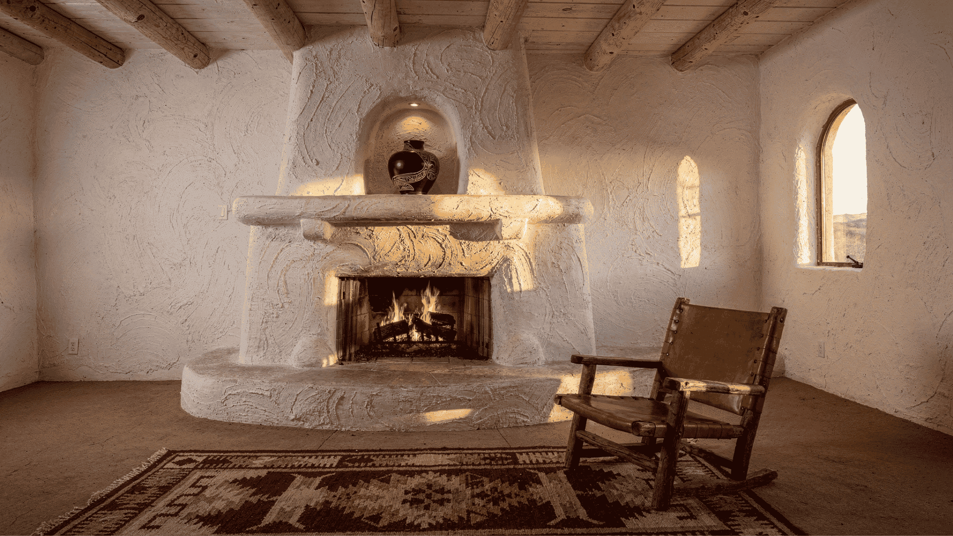 _a southwestern pueblo style room with a large hand applied white stucco kiva fireplace with a rounded curved form a built in niche holding a ceramic vessel and a raised stone hearth beside a wood