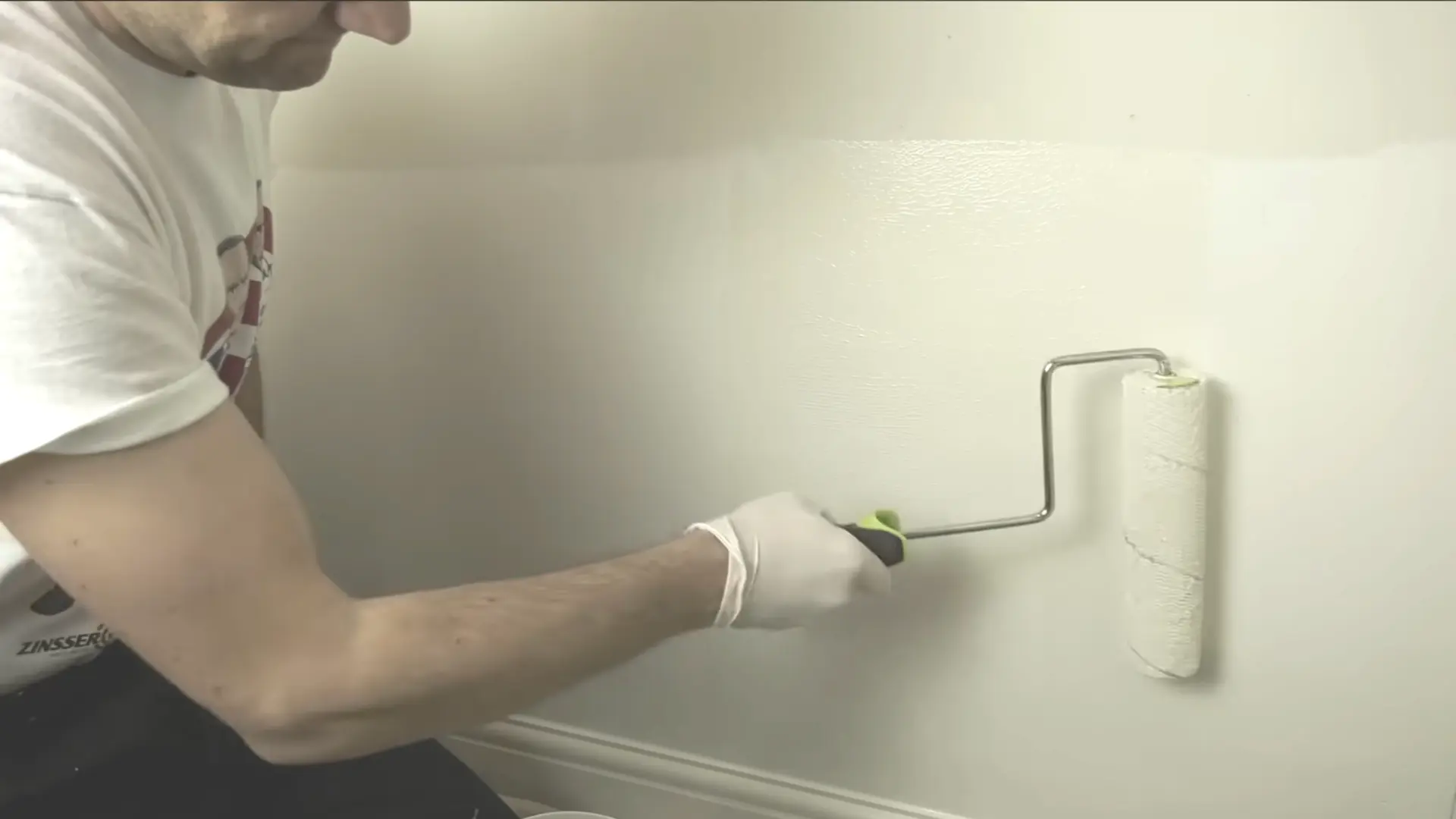 man in white tshirt with a rubber glove on his hand using a rolling pin to apply white paint on the wall