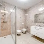 Modern bathroom with glass shower enclosure, white fixtures, and beige stone tile walls and floor