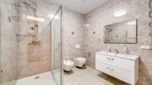 Modern bathroom with glass shower enclosure, white fixtures, and beige stone tile walls and floor