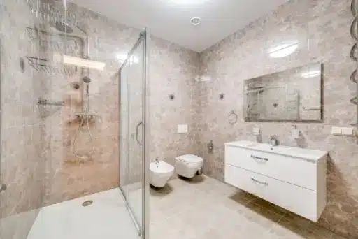 Modern bathroom with glass shower enclosure, white fixtures, and beige stone tile walls and floor