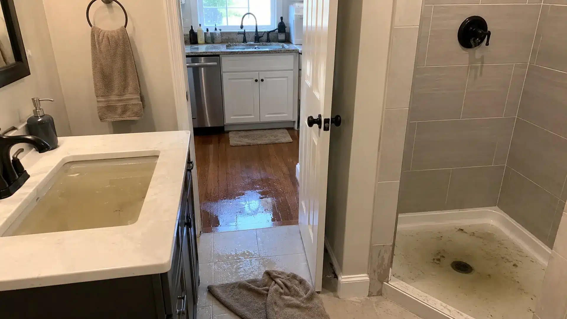 bathroom sink and shower filled with dirty backup water while water spreads across nearby hallway floor