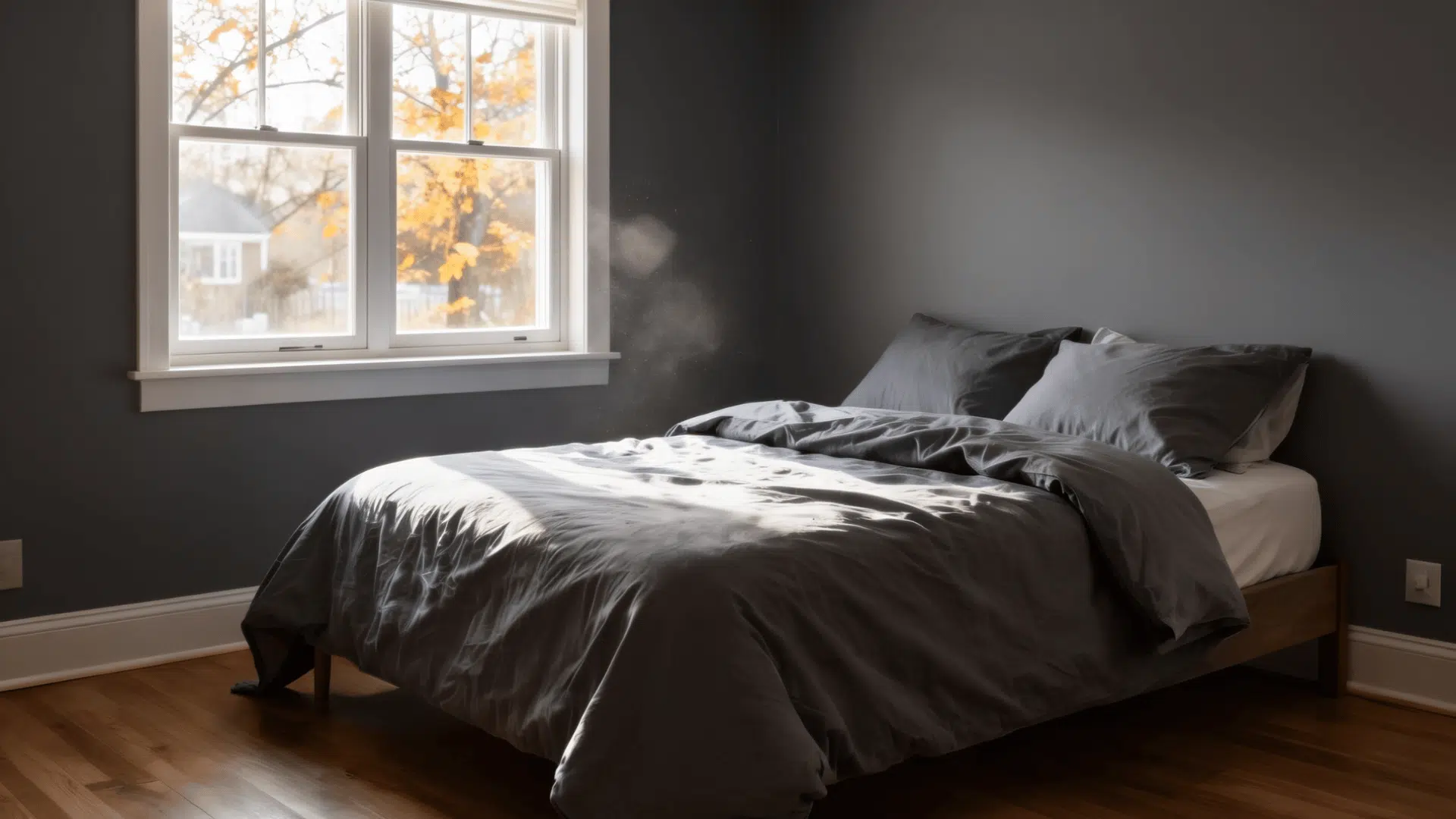 bed with gray bedding is positioned next to a window with autumn trees visible outside