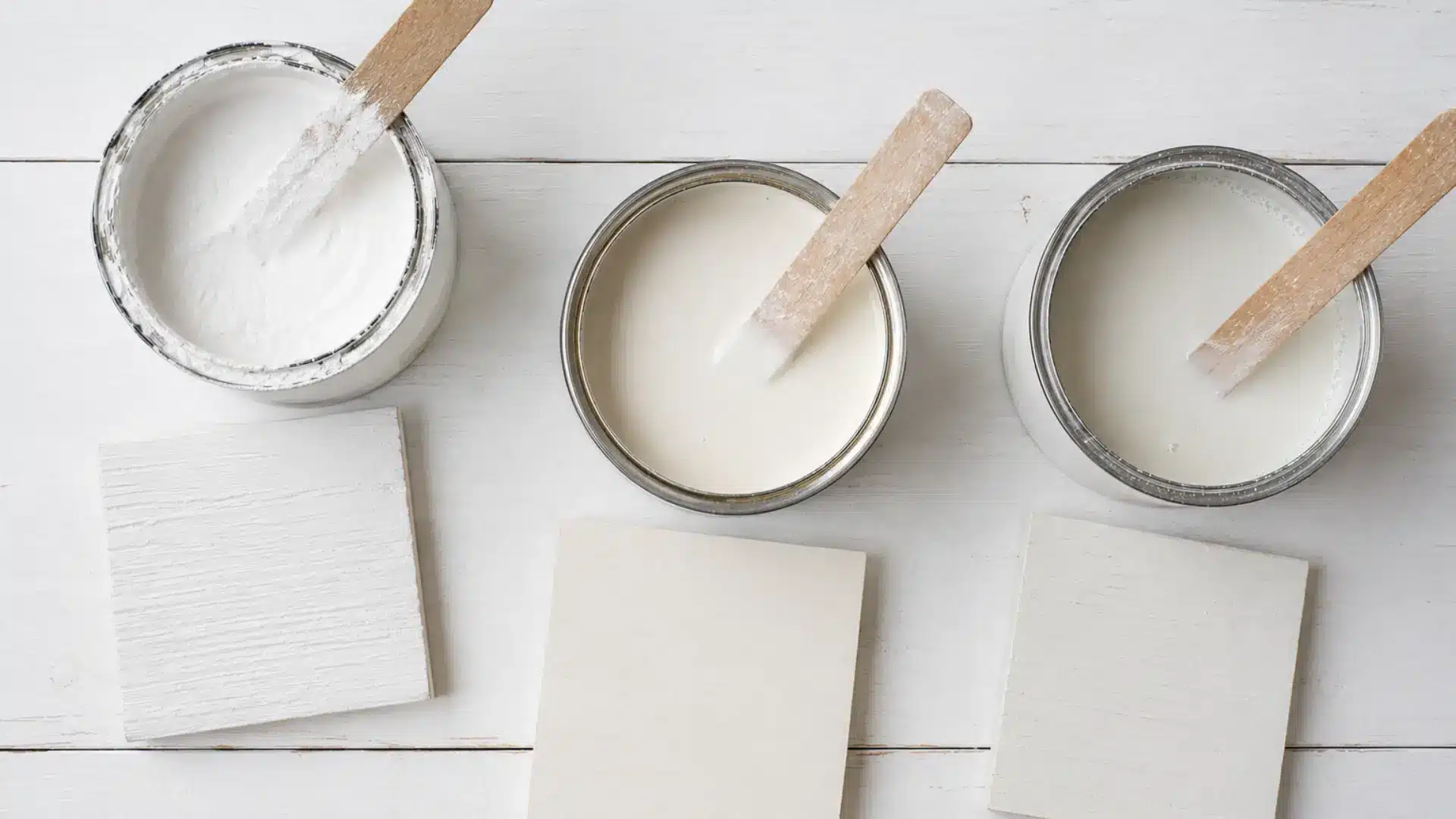 a clean top-down flat lay on a white wooden surface showing three small open paint tins arranged in a loose triangle, the left containing thick chalky matte white paint with a dry brushed rim, the center containing smooth creamy latex paint with a slightly shiny surface, and the right containing thin watery milk paint in a soft off-white tone, each with a wooden stir stick resting across it and a corresponding painted wood sample square placed beside it, all under soft even natural light
