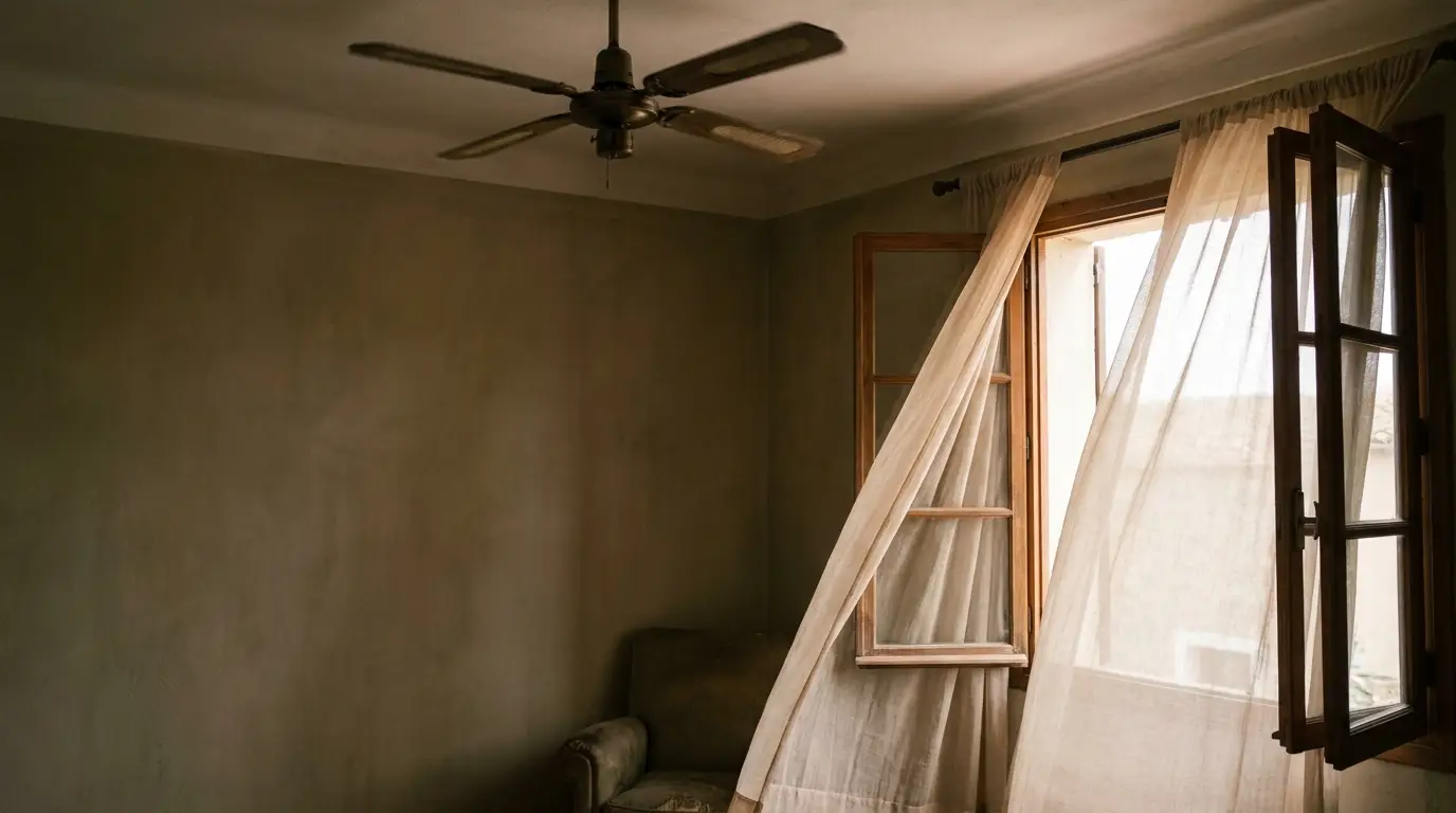Curtain billowing through open window in dimly lit room with ceiling fan and armchair