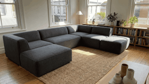 dark gray modular sectional sofa on a woven rug in a bright living room with large windows bookshelves and indoor plants