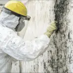 man in safety hazmat suit wearing a yellow helmet and face visor wiping off mold from a wall with a spatula