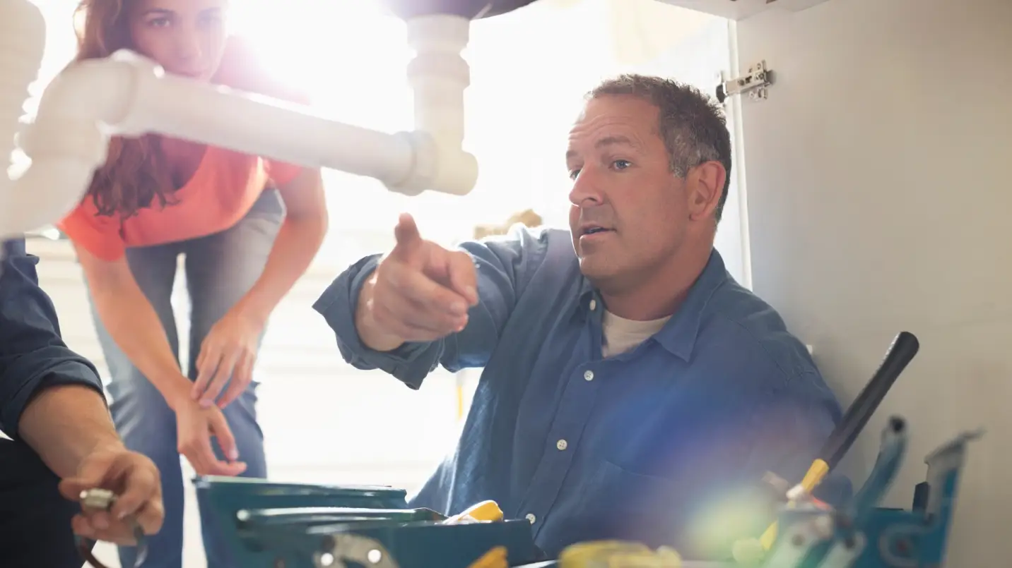 Man fixing plumbing under sink with woman observing in a brightly lit kitchen