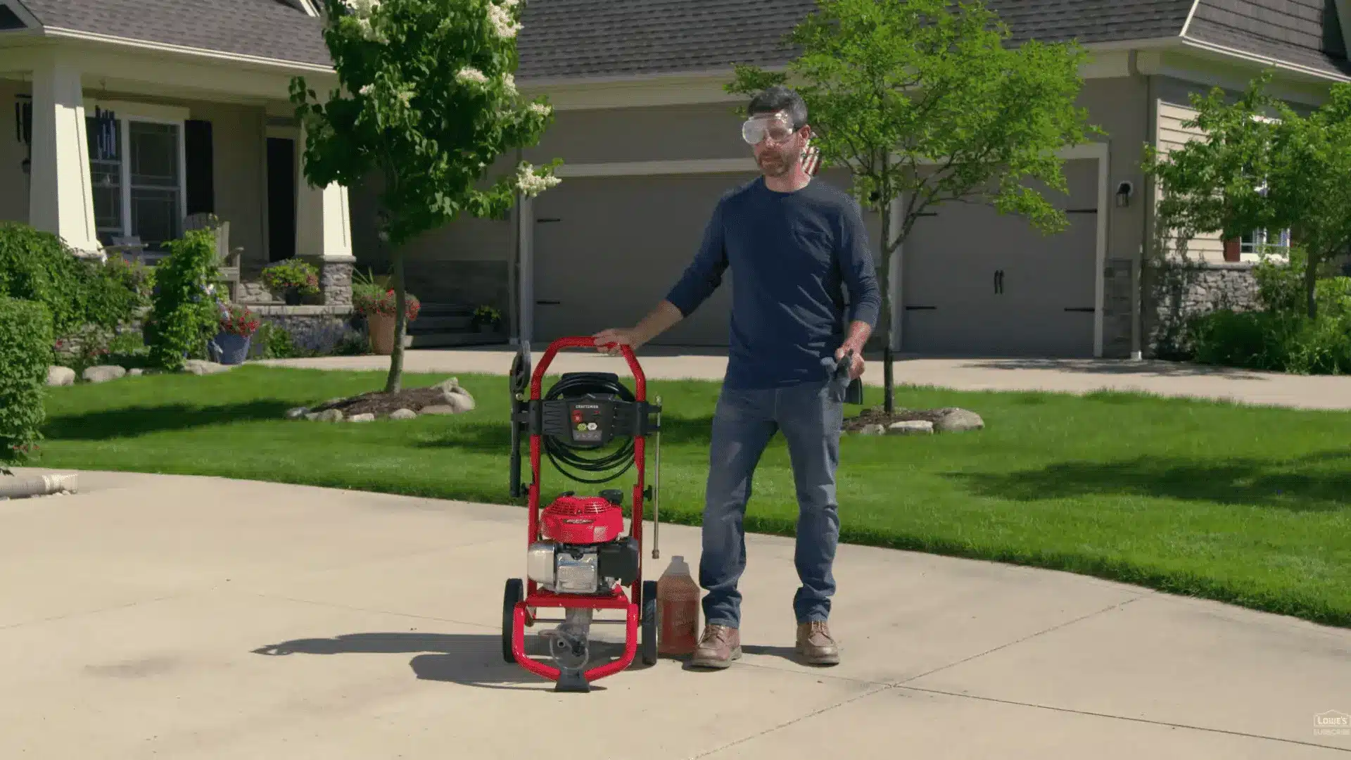 man wearing safety goggles and gloves standing beside a pressure washer in a driveway