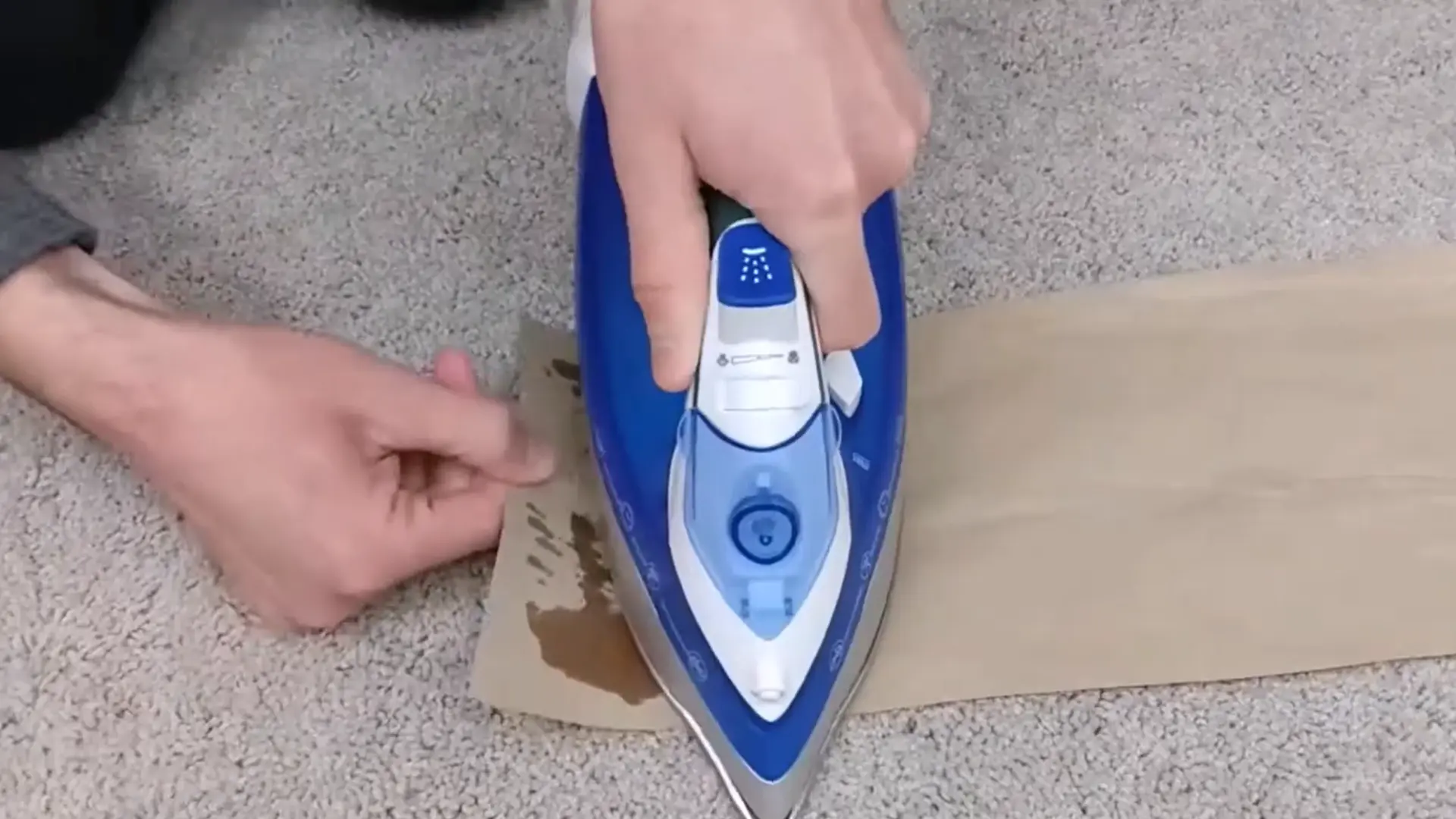 hands using a blue iron over brown paper to lift melted candle wax from gray carpet fibers