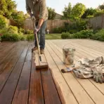 Homeowner staining a clean wooden deck with a brush, stain pad, bucket, and safety gloves