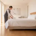 Housekeeper arranging towels on bed in bright, modern hotel room with wooden floor and bedside plant