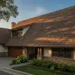 Large stone house with brown shingle roof and lush landscaping in evening light