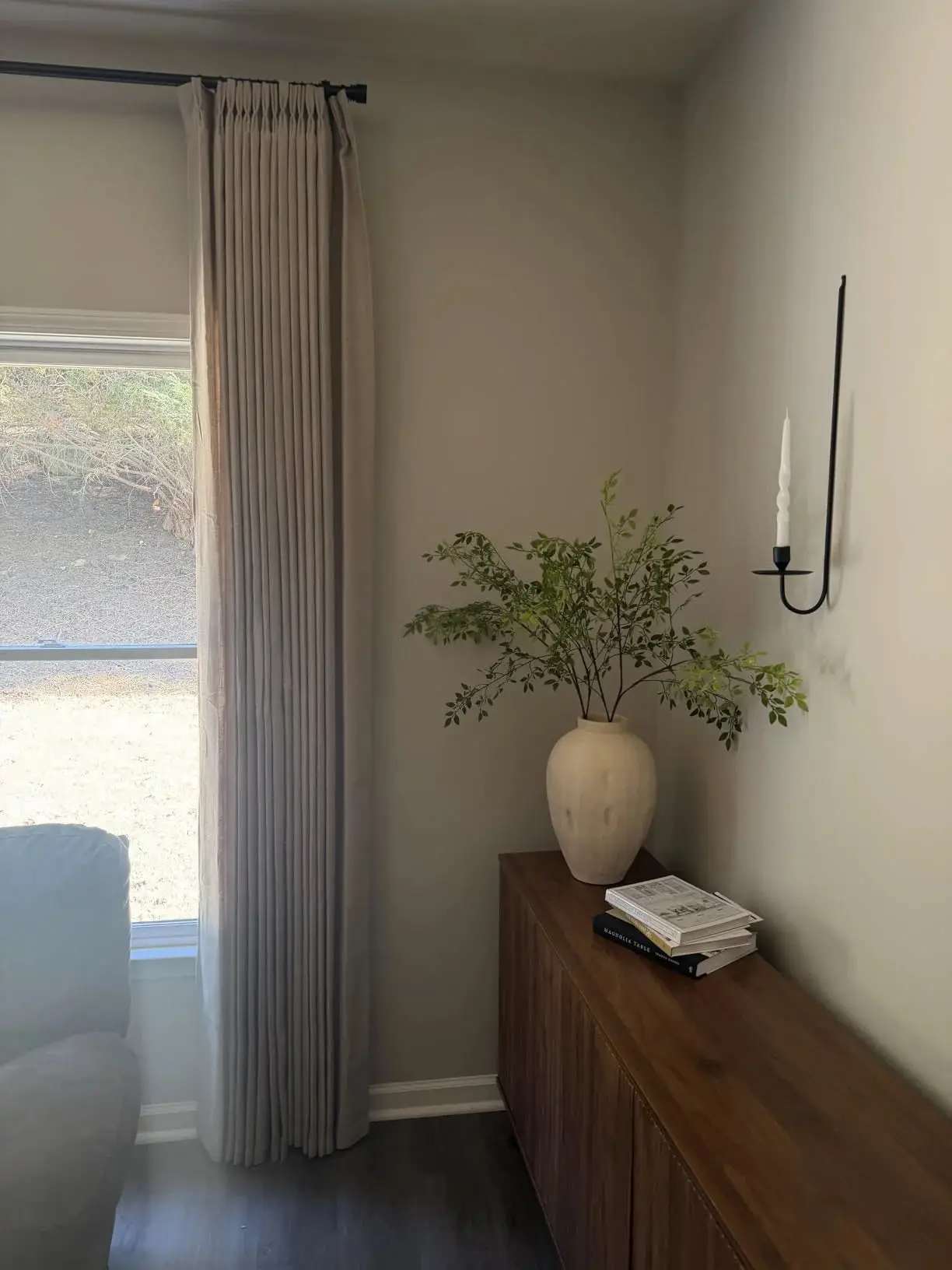 Vase with green branches on wooden sideboard beside tall curtains in living room corner