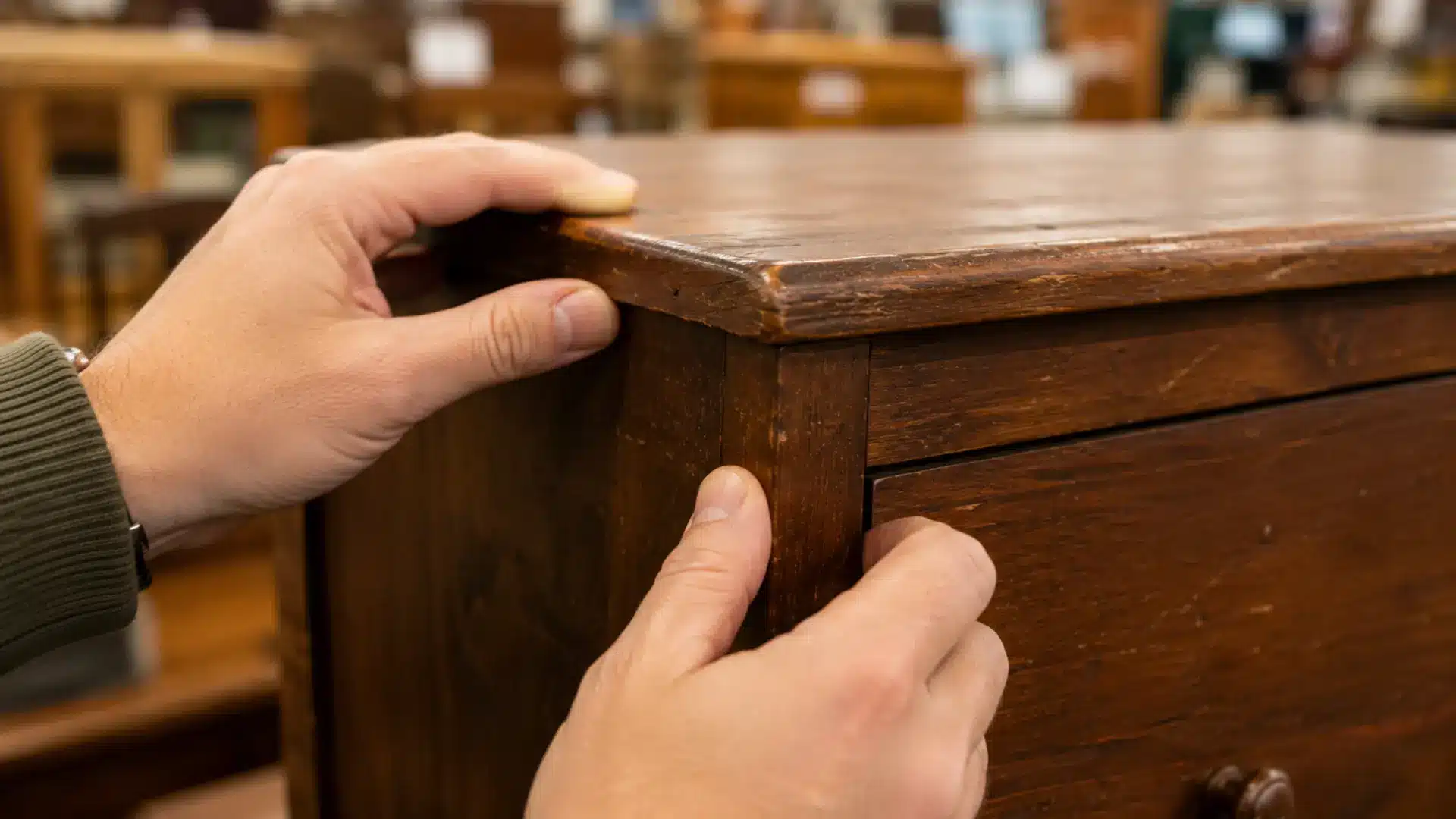 a close-up realistic photograph of a person's hands carefully inspecting the corner joint of a solid wooden dresser inside a thrift store, with one hand gripping the frame and the other pressing gently on the joint to test its sturdiness, the dresser showing an aged brown finish with minor scratches, and blurred shelves and other furniture pieces softly out of focus in the background under warm ambient thrift store lighting