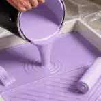 light purple enamel paint pouring into a tray with brush and roller on a protected work surface