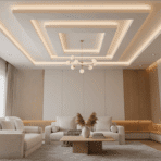 Modern living room with layered false ceiling and soft cove lighting. Neutral color scheme with white, beige, and light wood tones.