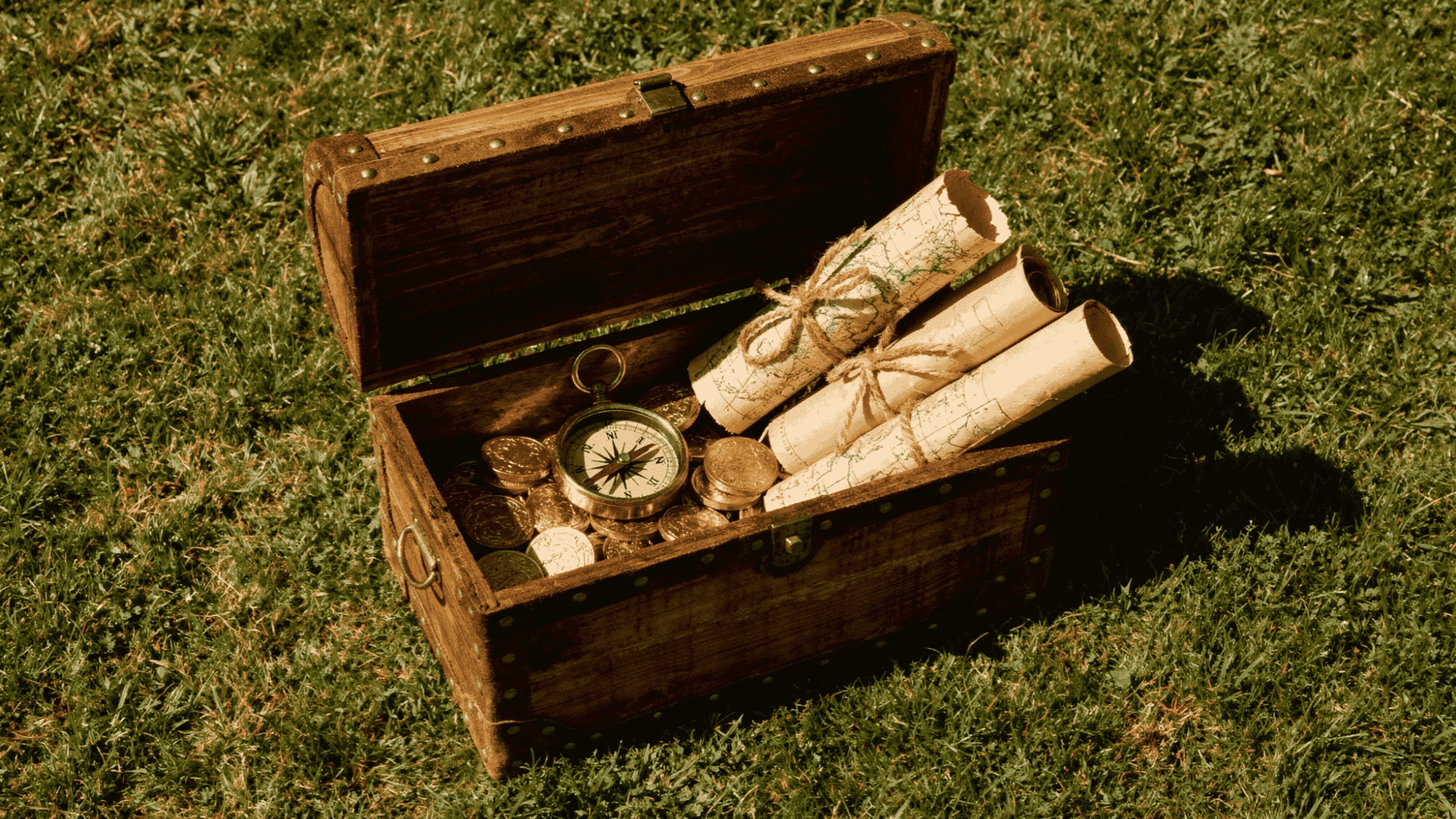 _open wooden treasure chest with rolled maps, gold coins, and a compass on grass