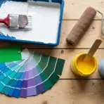 Paint supplies including a color swatch fan deck, paint roller, brush in a tray of white paint, and open jars of yellow and blue paint resting on a wooden surface