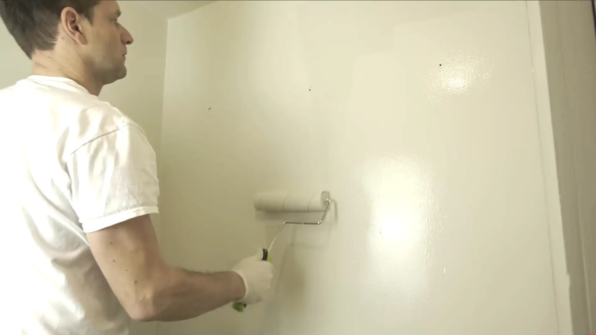 man in a white tshirt applying the second coat of white paint with a rolling pin brush on the wall