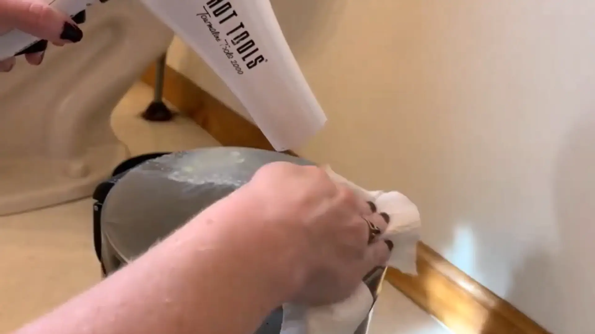 person using a hair dryer and cloth to remove soft candle wax from a metal surface indoors