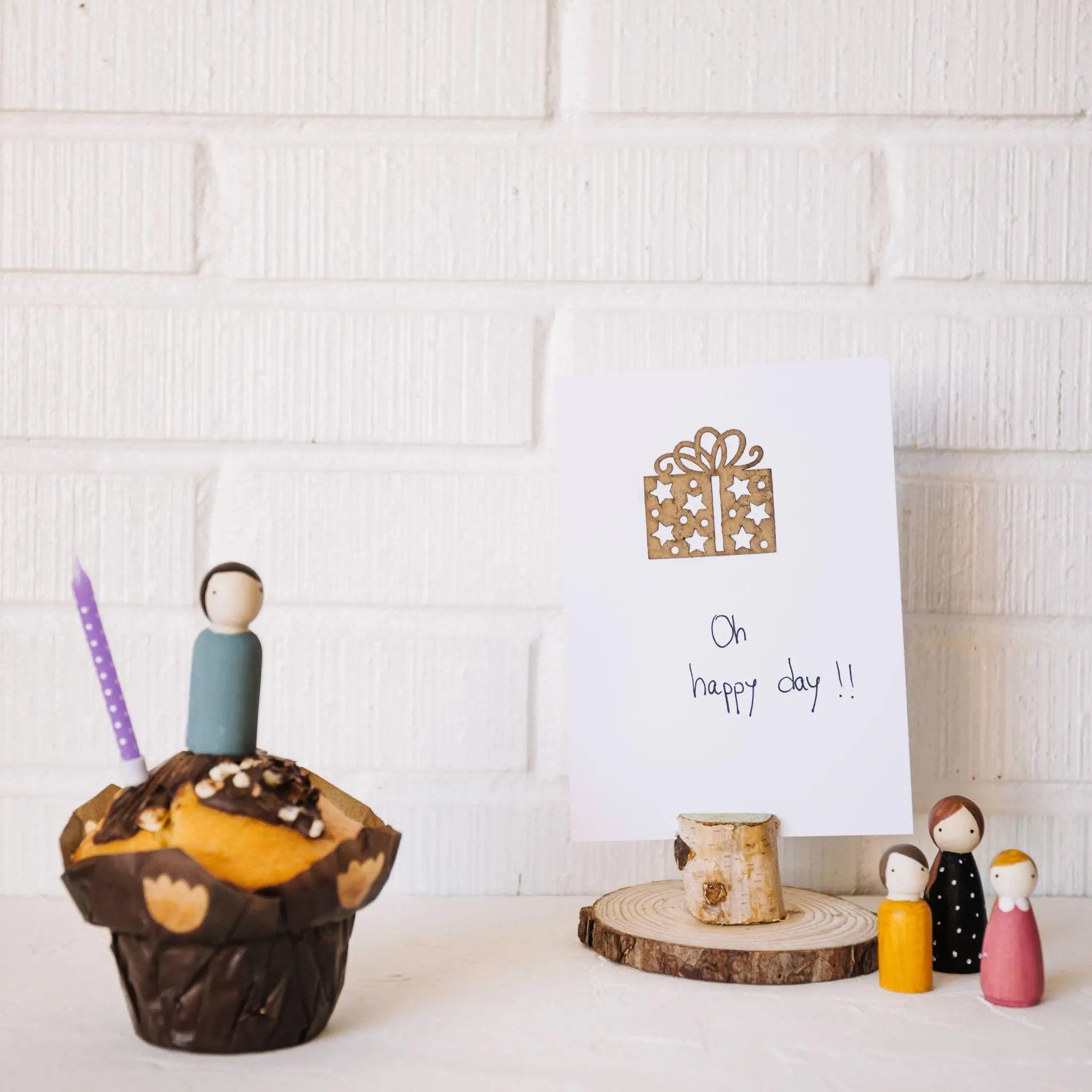 Cupcake with decorative topper and purple candle beside greeting card on white brick background