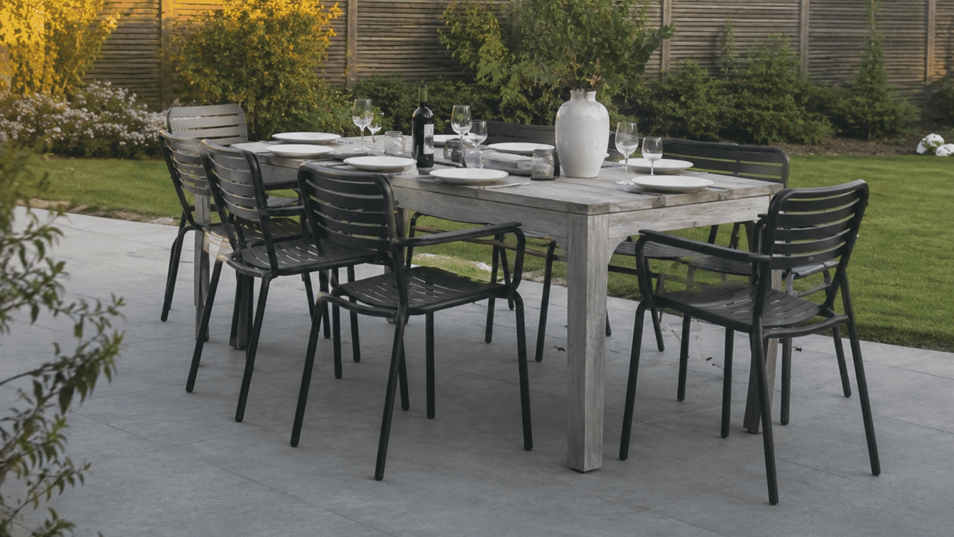 realistic outdoor dining table setup with black chairs on modern patio surrounded by green backyard garden