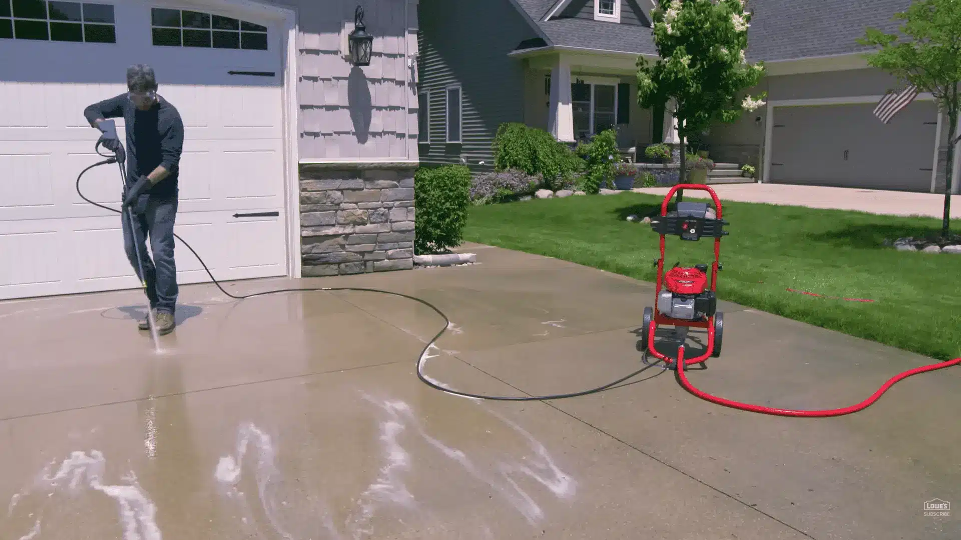 man pressure washing a concrete driveway with a red pressure washer and hose stretched across the yard