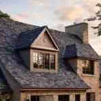 large stone house with natural slate roof featuring dormer window and stone chimney at dusk