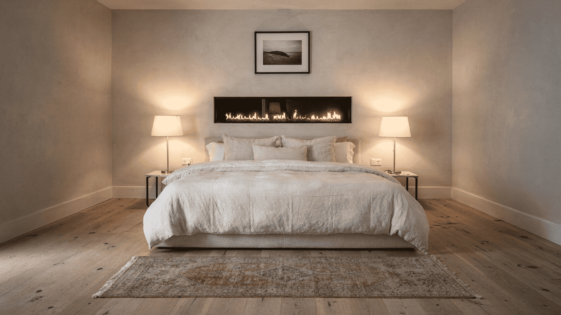 slim linear gas fireplace mounted into the wall above the bed head flanked by matching table lamps on a plaster wall with linen bedding and a vintage rug on wide plank floorboards