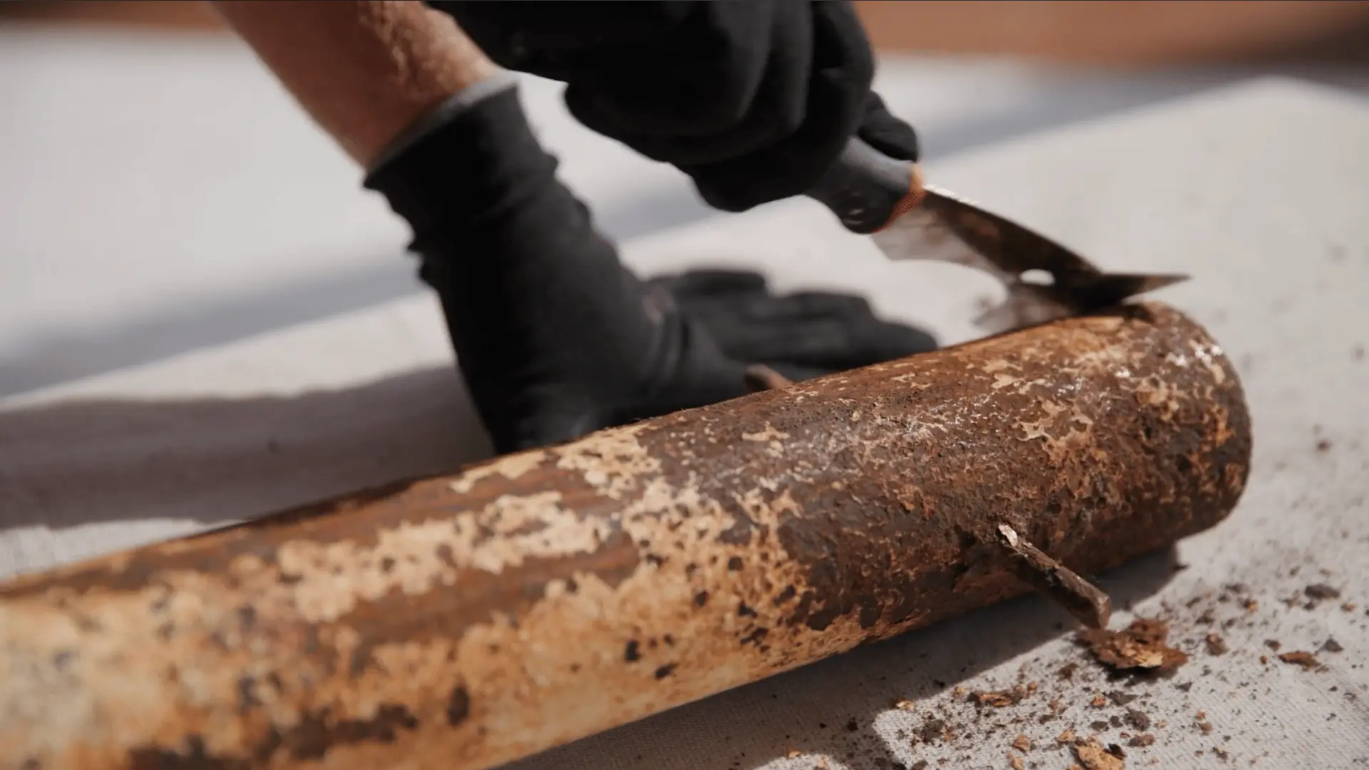 gloved hand using a paint scraper to remove peeling paint and rust from a metal pipe before repainting