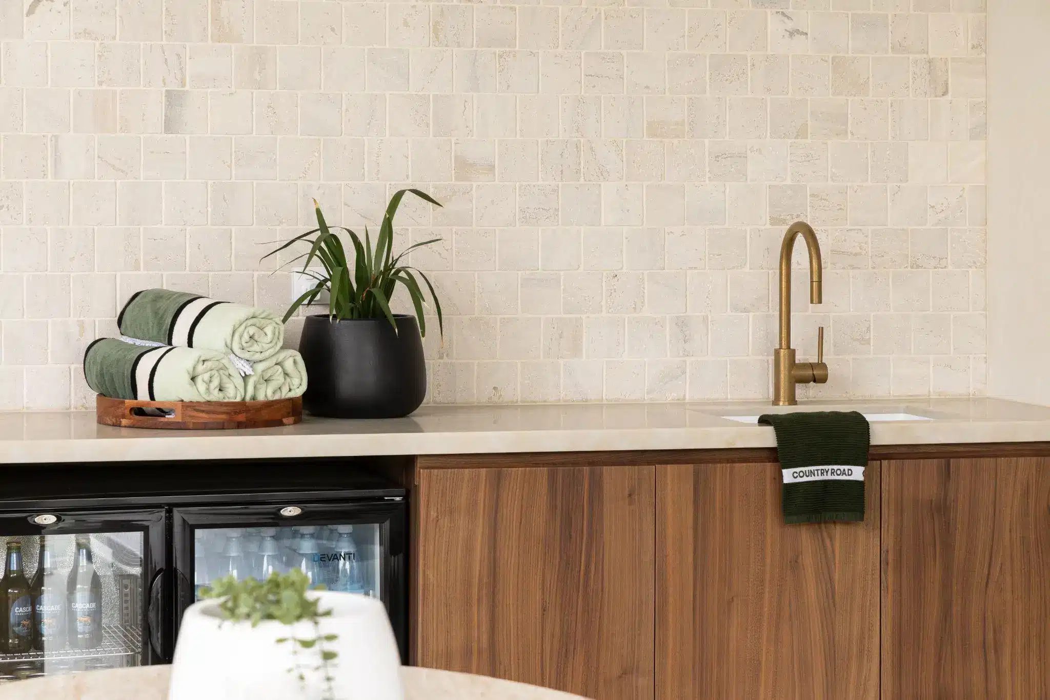 Countertop with green rolled towels and plant beside brass faucet and wooden cabinets