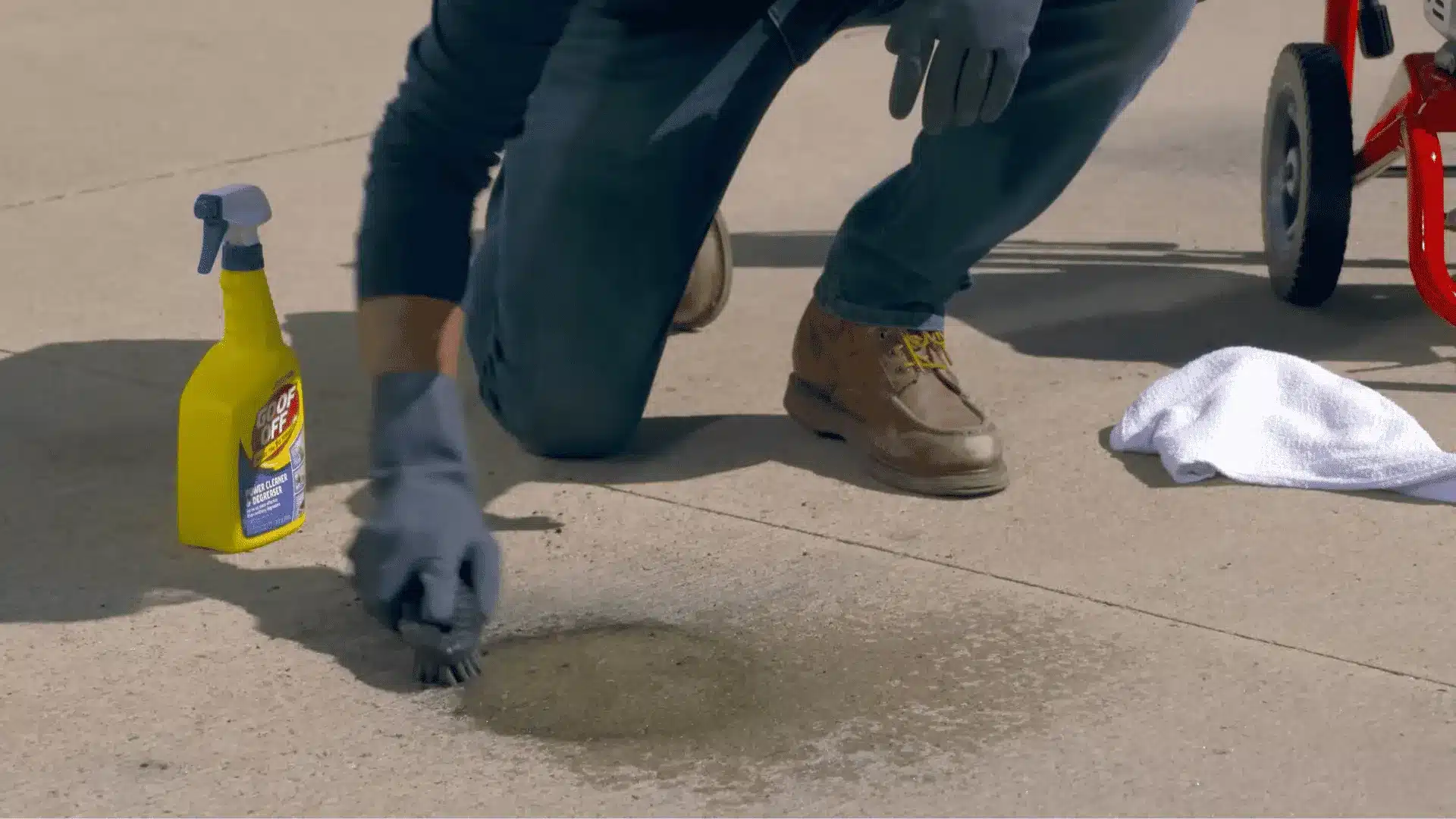 man in gloves scrubbing a dark oil stain on a driveway with a brush and degreaser spray bottle nearby