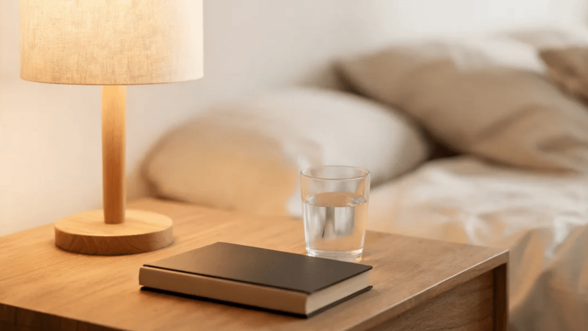 wooden nightstand with lamp glass of water and book next to beige bed