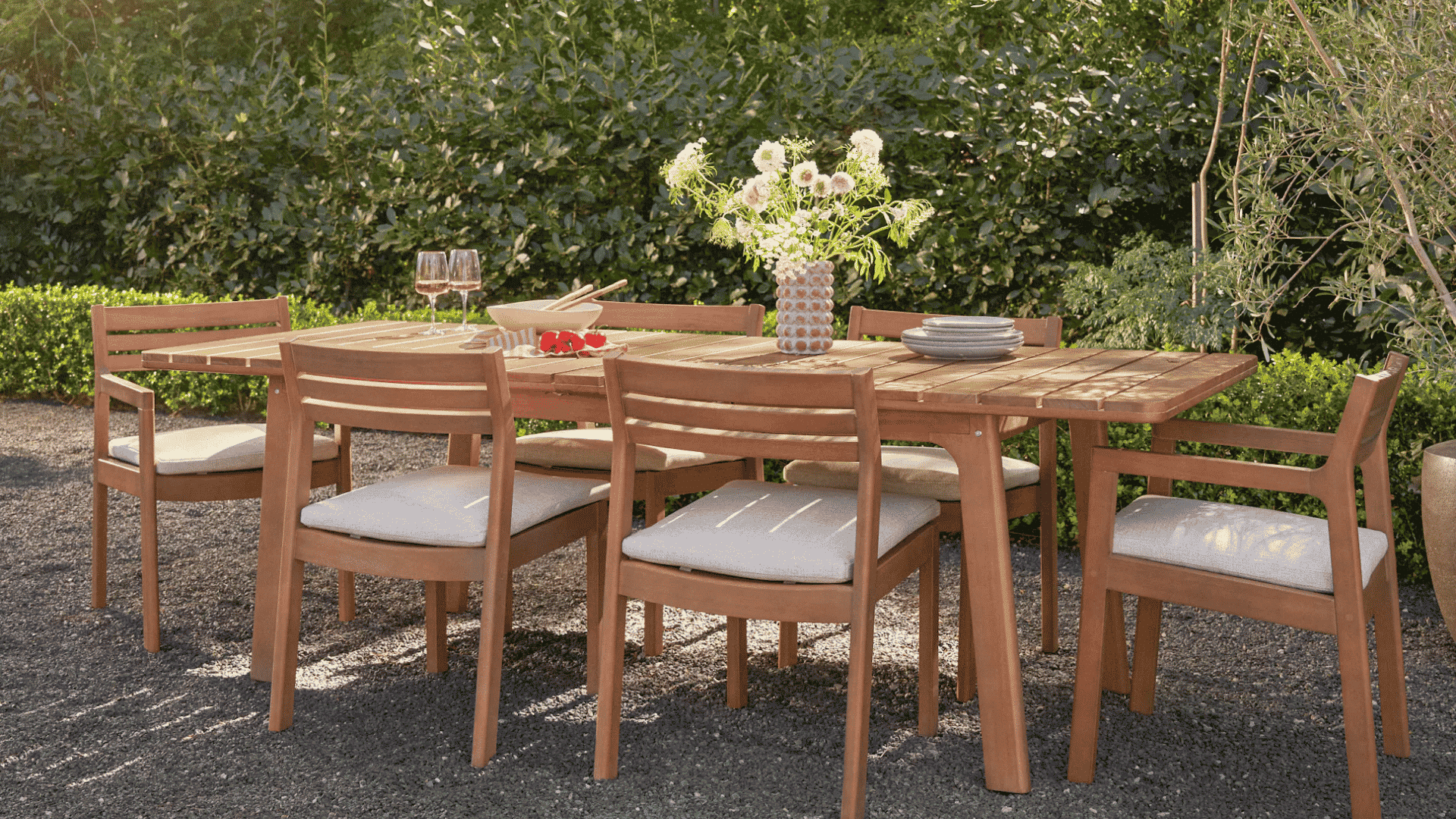 wooden outdoor dining set with cushioned chairs placed on gravel patio surrounded by lush greenery