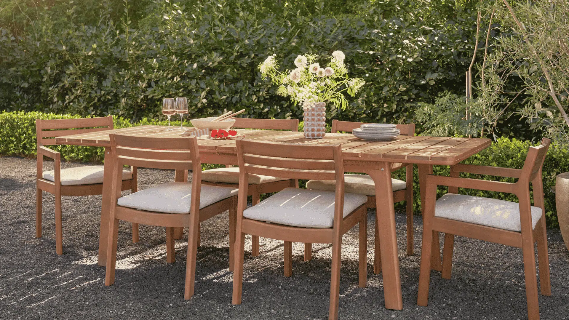 wooden outdoor dining set with cushioned chairs placed on gravel patio surrounded by lush greenery