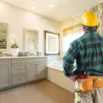 Construction worker in plaid shirt and tool belt inspecting modern bathroom with tile backsplash