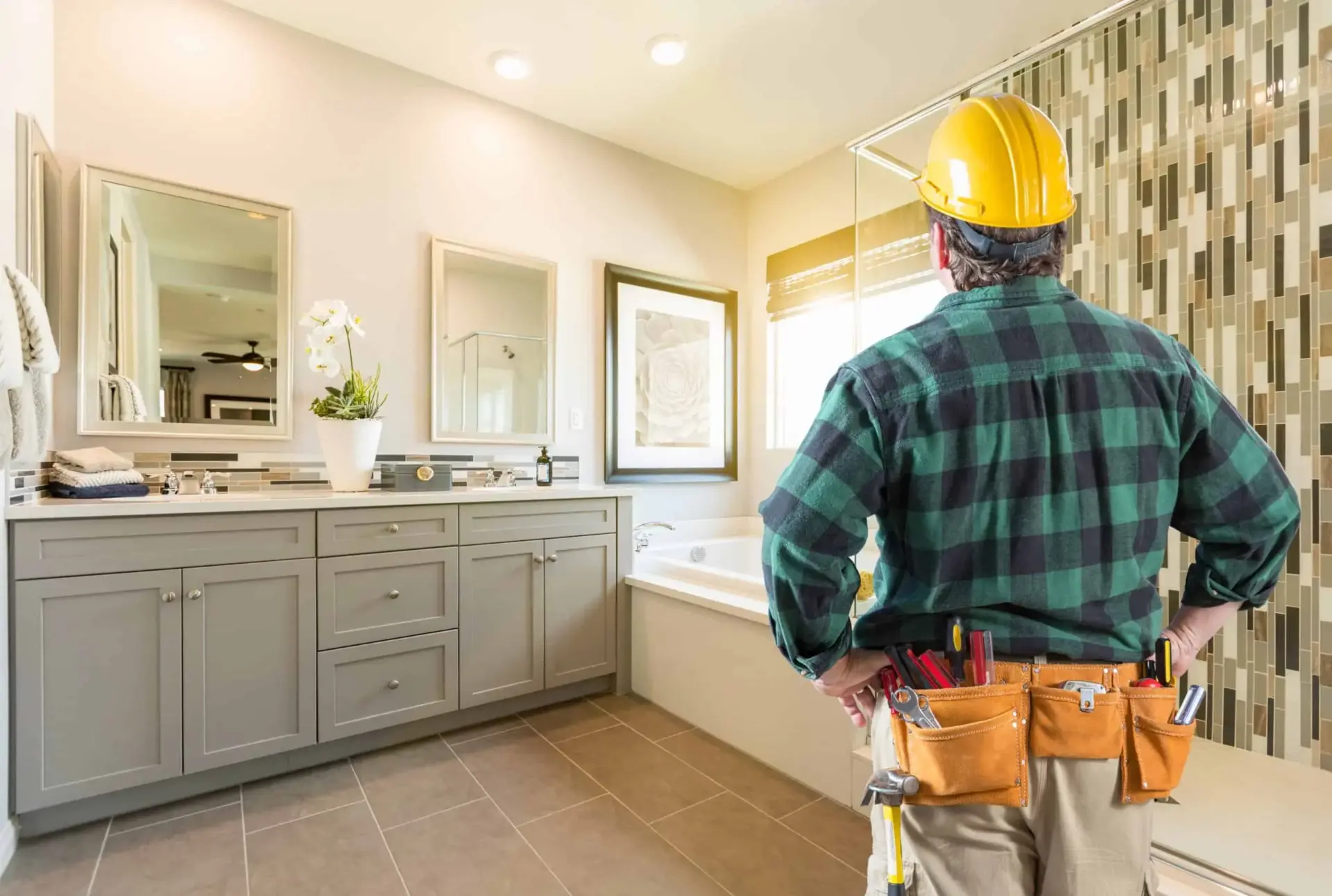 Construction worker in plaid shirt and tool belt inspecting modern bathroom with tile backsplash