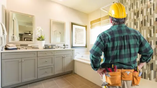 Construction worker in plaid shirt and tool belt inspecting modern bathroom with tile backsplash