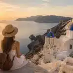 girl sitting above a coastal village with white buildings, blue domes and sunset sea views