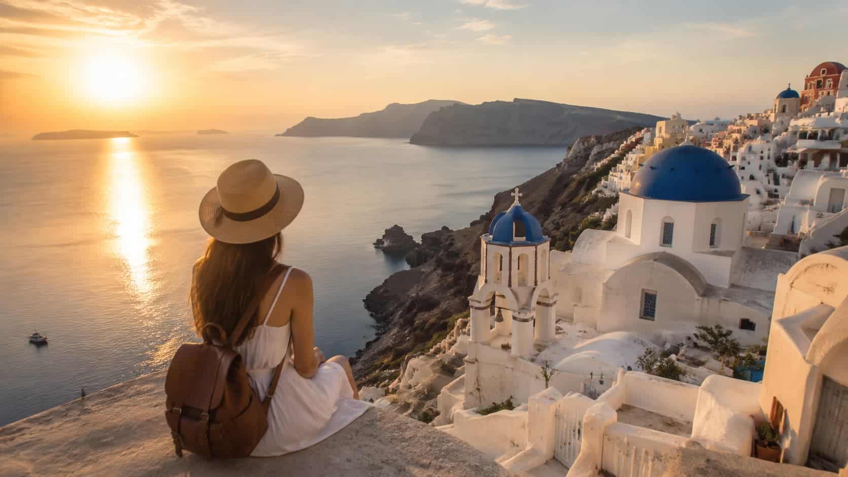 girl sitting above a coastal village with white buildings, blue domes and sunset sea views