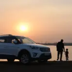 White SUV parked by lakeside at sunset with man and child walking nearby
