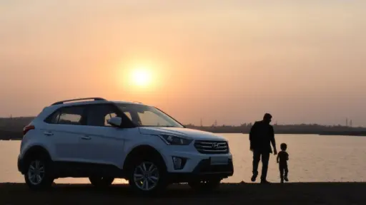 White SUV parked by lakeside at sunset with man and child walking nearby