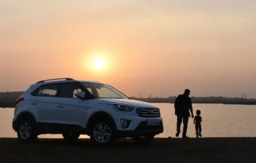 White SUV parked by lakeside at sunset with man and child walking nearby