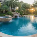 Luxury backyard pool with waterfall feature surrounded by palm trees at sunset