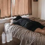 person lying on a bed with closed curtains frozen water bottles and a standing fan cooling the room