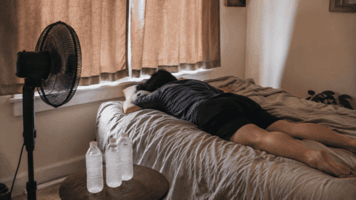 person lying on a bed with closed curtains frozen water bottles and a standing fan cooling the room